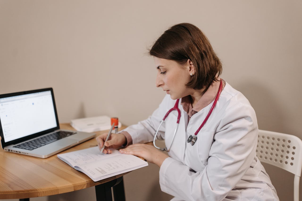 services-05 A female doctor in a lab coat writing a prescription in a clinic office setting with a laptop on the table.