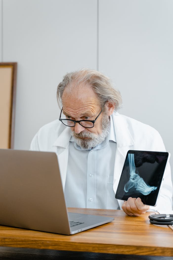 why-choose-us Senior doctor analyzing a foot X-ray while working on a laptop in a medical office.